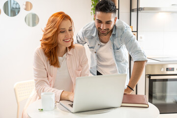 Wall Mural - Beautiful ginger young woman and middle eastern young man using laptop at home. Diverse students working homework together.