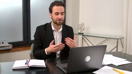 Wall Mural - Smiling young businessman in formal wear sitting at the desk in office, studying or working online, having virtual meeting, waving at the laptop screen, male employee takes a part in online conference