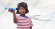 African american boy with toy plane wearing aviator hat and goggles and airplane flying against sky