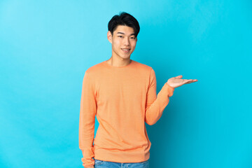 Wall Mural - Young Chinese man isolated on blue background holding copyspace imaginary on the palm to insert an ad