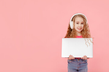 smiling adorable child girl is holding laptop and in headphones on pink background. study education concept