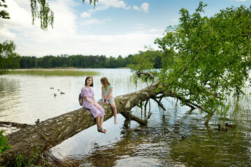 Wall Mural - Two cute young girls sitting on a faller tree by the river or lake dipping their feet in the water on warm summer day.