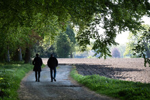 Unrecognizable Man And Woman Walking On A Sandy Path Under A Canopy Of Trees Past A Forest And A Farm Field