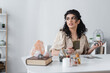 © LIGHTFIELD STUDIOS - Smiling gypsy medium holding tarot cards near blurred book and crystals on table.