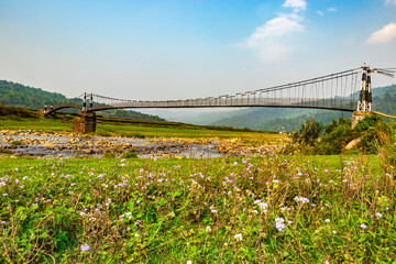 isolated iron suspension bridge over flowing river with mountain and blue sky background at morning