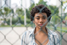 Close-up Of A Young Latin Woman Looking At The Camera With Serious Face While Standing Behind Woven Wire Fence At The Park.
