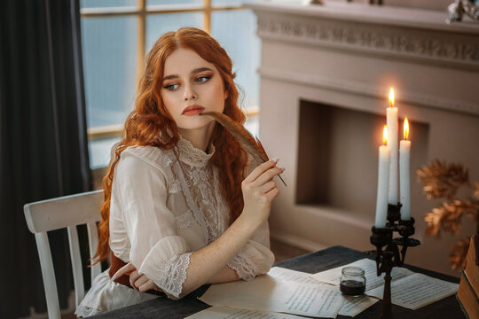 Portrait red-haired vintage woman writer holding bird feather in hands. Medieval girl sit at table writes book, letters on sheets paper with ink pen. Photo old retro style. White blouse, classic room