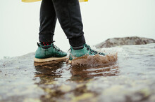 Male Tourist Standing On A Puddle On A Rock On A Background Of Misty Views, Photo Of Feet In Green Boots In The Water.