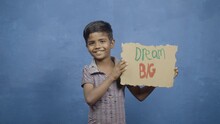 Happy Smiling Kid With Dream Big Sign Board Standing Against Blue Background By Looking At Camera - Concept Of Motivation, Inspirational And Childhood Aspirations.