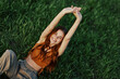 © SHOTPRIME STUDIO - A woman lying in nature on the green grass in the park resting with her hands up and smiling with her mouth open, her red hair flying in the wind in the open air in the sunlight. Freelancer's concept