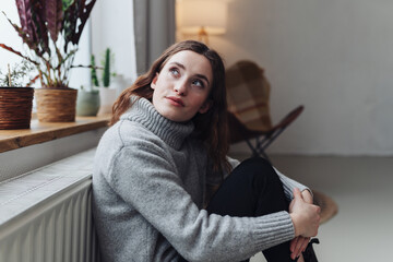 Young woman sitting on the floor leaning against a heater and looking out the window