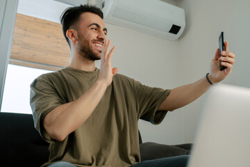 Wall Mural - Video chat, young handsome man in the living room video chatting with his close friend, smiling adult male waving at the phone