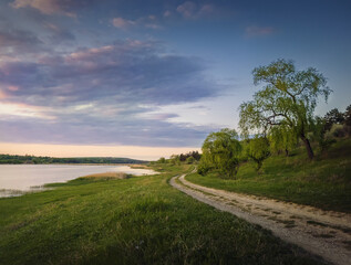 Rural landscape with a country road between lake and forest. Idyllic summer scene, peaceful evening with colorful sunset sky