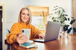 © baranq - Smiling mature woman using smartphone working at home office with laptop sitting at dinning table