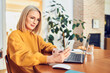 © baranq - Mature woman with glasses looking on smartphone sitting at dinning table while working from home