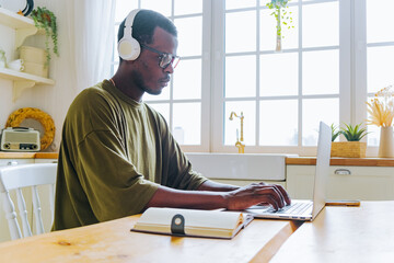 African American man in headphones takes online courses via laptop writing down notes. Guy listens to audio at table against big window