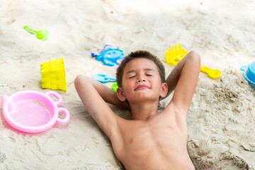 Little cute Asian child boy laying on tropical beach playing sand and beach toy with smiling on summer vacation. Happy children kid enjoy and fun outdoor activity lifestyle on beach holiday travel