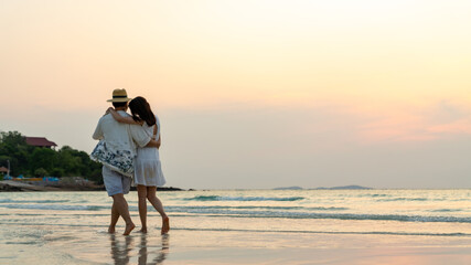 Asian couple holding hands and embracing each other while walking on tropical beach together at summer sunset. Happy family husband and wife enjoy outdoor lifestyle on holiday travel beach vacation