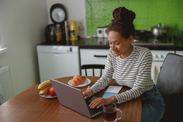 young positive woman working on computer sitting at table in kitchen in good mood