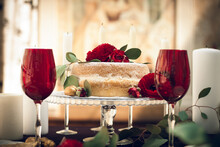 A Banquet Table In A Restaurant, With Red Wine Glasses, A Stylish Cake On A Glass Tray, Decorated With Red Flowers, Green Leaves, Candles And Golden Walnuts, For The Newlyweds.