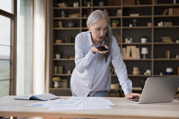 Wall Mural - Serious busy mature business professional woman standing at table with paper marketing reports, recording audio message on smartphone, using work online app on laptop computer, speaking on cell