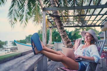 Wall Mural - Technology and travel. Working outdoors. Freelance concept. Pretty young woman using laptop in cafe on tropical beach.