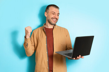 Poster - Joyful Man Using Laptop Shaking Fists On Blue Background