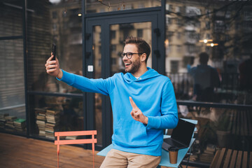 Wall Mural - Happy Caucasian man using front mobile camera on modern cellular technology for making selfie content during weekend leisure, smiling male millennial shooting video vlog via smartphone application