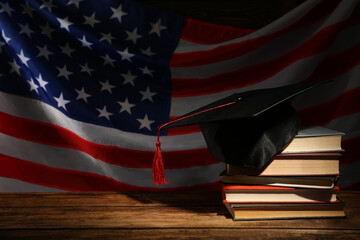 Wall Mural - Graduation hat and books on wooden table against American flag in darkness, space for text