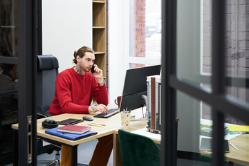Poster - Serious young manager having a conversation on mobile phone while working on computer at his workplace at office