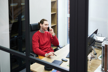 Poster - Smiling manager sitting at table at office in front of computer monitor and having business conversation on mobile phone