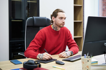 Poster - Young designer sitting at desk in front of computer monitor and using graphic tablet to do his work
