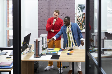 Poster - African young architect pointing at mock-up of buildings on table and talking to his colleague