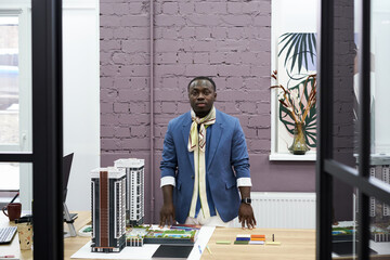 Poster - Portrait of African professional architect looking at camera while standing at table with his project of new buildings