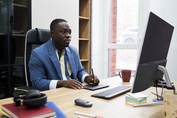 Poster - Serious African designer looking at computer monitor while working on graphic tablet at table at office