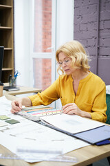 Poster - Serious female designer in eyeglasses sitting at table and examining colours in palette for future project