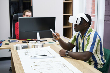 Poster - African young architect using virtual reality headset to look at construction project while sitting at table with blueprint