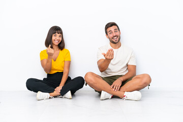 Wall Mural - Young couple sitting on the floor isolated on white background inviting to come with hand. Happy that you came