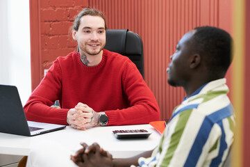 Poster - Young smiling businessman sitting at table with laptop and talking to client during their meeting at office