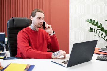 Poster - Young businessman sitting at office desk with laptop and having conversation on mobile phone