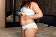 © Юля Бурмистрова - Curly plus size woman in white underwear standing near bed in bedroom and touching stomach with stretch marks closeup side view. Fat burning treatment of thick belly. Weight loss program, dieting.