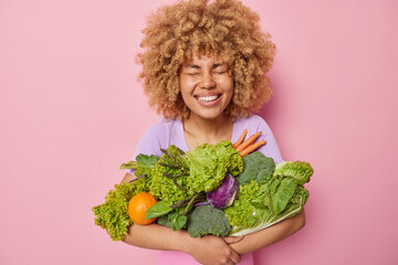 Wall Mural - Horizontal shot of cheerful happy young woman with curly hair embraces big bouquet of green vegetables glad to buy groceries at market smiles toothily isolated over pink background. Agriculture