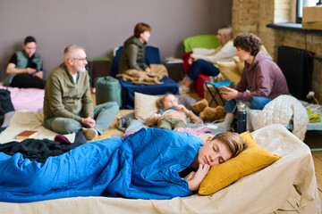 Wall Mural - Adolescent boy keeping his head on pillow while sleeping under blue blanket against group of people communicating on their beds