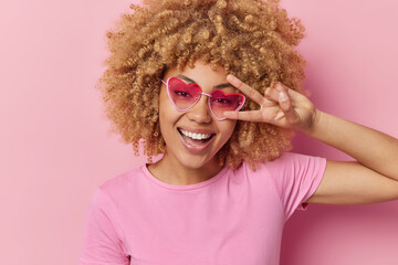 Wall Mural - Portrait of happy curly haired woman makes peace gesture over eye smiles toothily has glad expression foolishes around wears heart sunglasses and casual t shirt isolated over pink background.