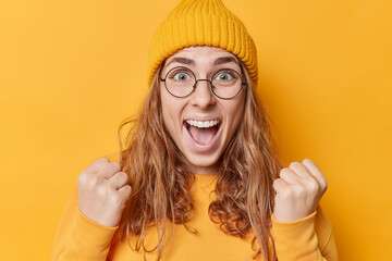 Big win concept. Joyful female winner celebrates victory clenches fists and yells loudly hooray feels euphoric wears round transparent eyeglasses hat casual sweatshirt isolated over yellow wall