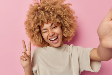 Wall Mural - Beautiful cheerful woman winks eye laughs positively makes peace gesture poses for selfie poses against pink background keeps arm outstreched stays positive has video chatting on smartphone app