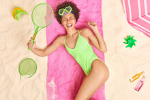 Overjoyed Curly Haired Woman Rests After Playing Active Game At Beach Holds Tennis Racket Laughs Gladfully Wears Green Swim Suit Lies On Pink Towel Surrounded By Different Items. Recreation Concept