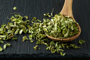 A closeup of a wooden spoon with moringa leaves scattered on a black slate