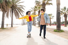 Happy Man With Woman Skateboarding In Front Of Palm Trees On Footpath