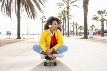 Smiling Woman Wearing Headphones Sitting On Skateboard In Front Of Palm Trees